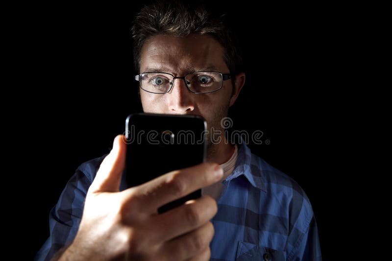 Close up portrait of young man looking intensively to mobile phone screen with blue eyes wide open isolated on black background. Close up portrait of young man stock photo