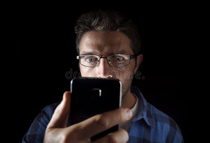 Close up portrait of young man looking intensively to mobile phone screen with blue eyes wide open isolated on black background. Close up portrait of young man royalty free stock image