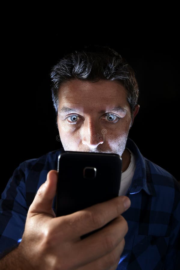 Close up portrait of young man looking intensively to mobile phone screen with blue eyes wide open isolated on black background. Close up portrait of young man stock photography